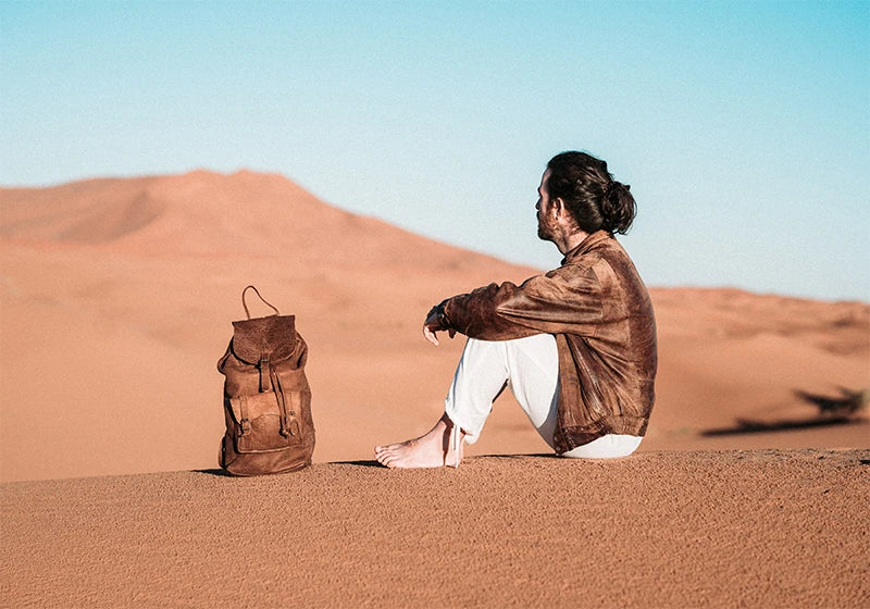 Man sitting on a desert dune beside a brown Rowdy leather rucksack