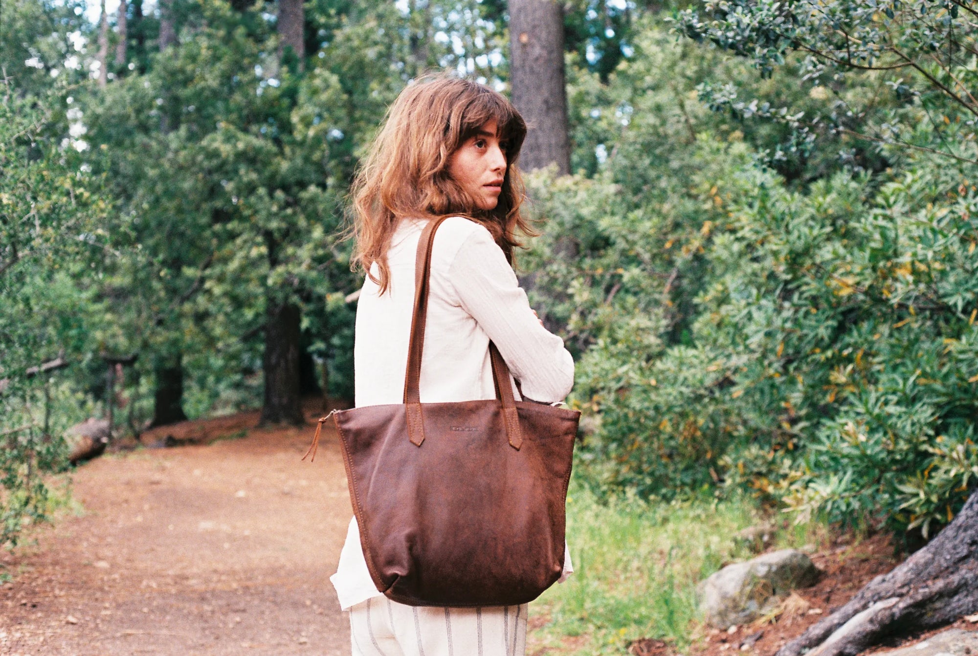 Woman on a forest path carrying a brown Rowdy leather tote on her shoulder